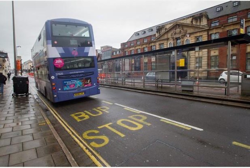 A bus on a street in Bristol pulling away from a Bus Stop. Instead of saying Bus Stop on the road it says Bup Stop. 
